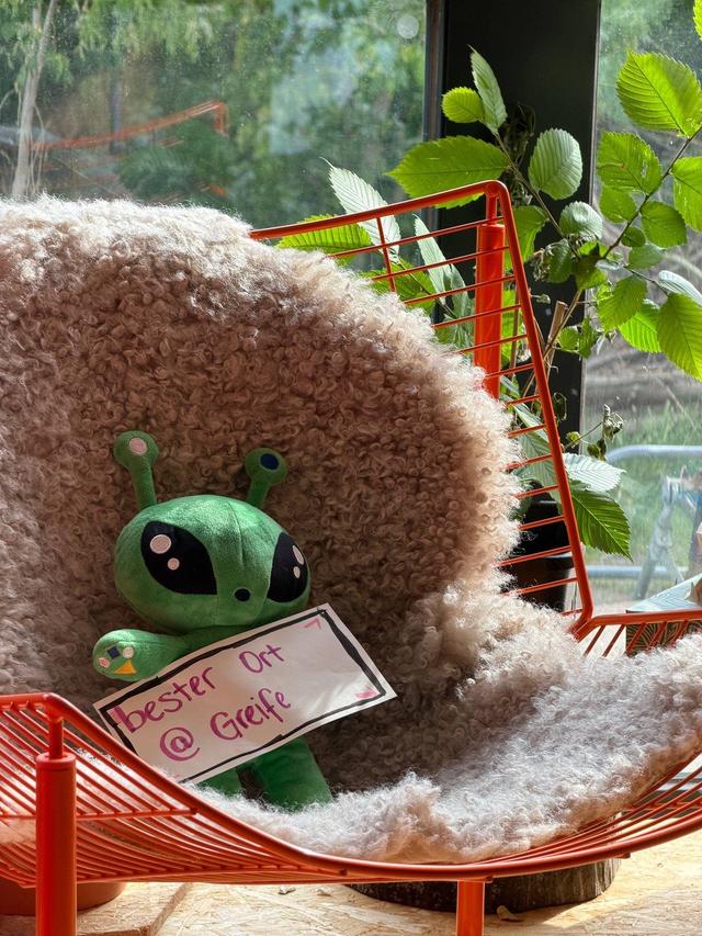 Grünes Plüsch-Alien mit dem handgeschriebenen Schild „bester Ort @ Greife“ in einem orangefarbenen Drahtsessel mit Schaffell, dahinter Grün vor dem Fenster