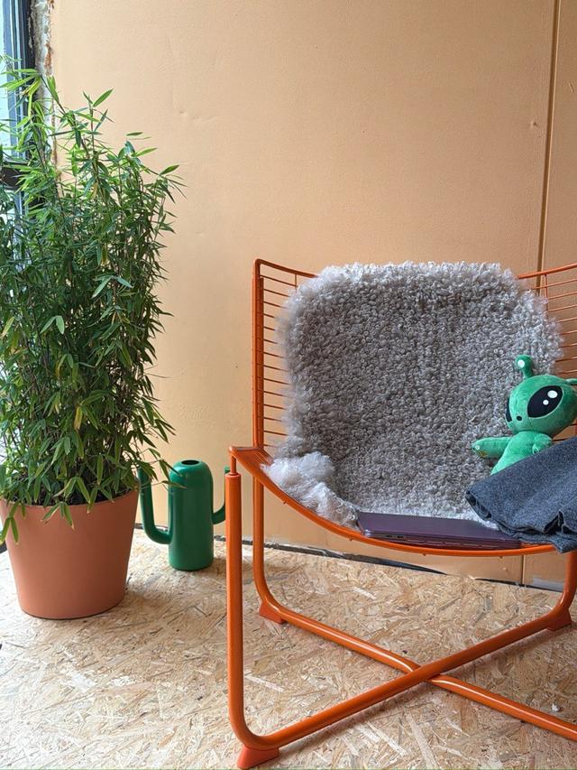 Sitzecke vor einer gelben Wand: orangefarbener Drahtsessel mit Schaffell und Plüsch-Alien, daneben ein Bambus im Topf