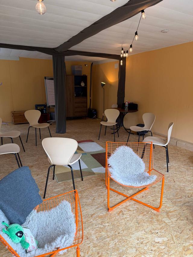 Stuhlkreis im Seminarraum: helle Holzstühle und zwei orangefarbene Drahtsessel um einen bunten Teppich, Lichterkette unter der Decke, hinten ein Flipchart