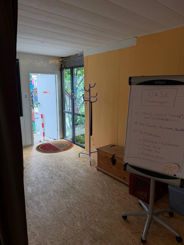 Eingang des Seminarraums mit Glastür, Fußmatte und Garderobenständer, rechts ein Flipchart mit handgeschriebenen Raumregeln