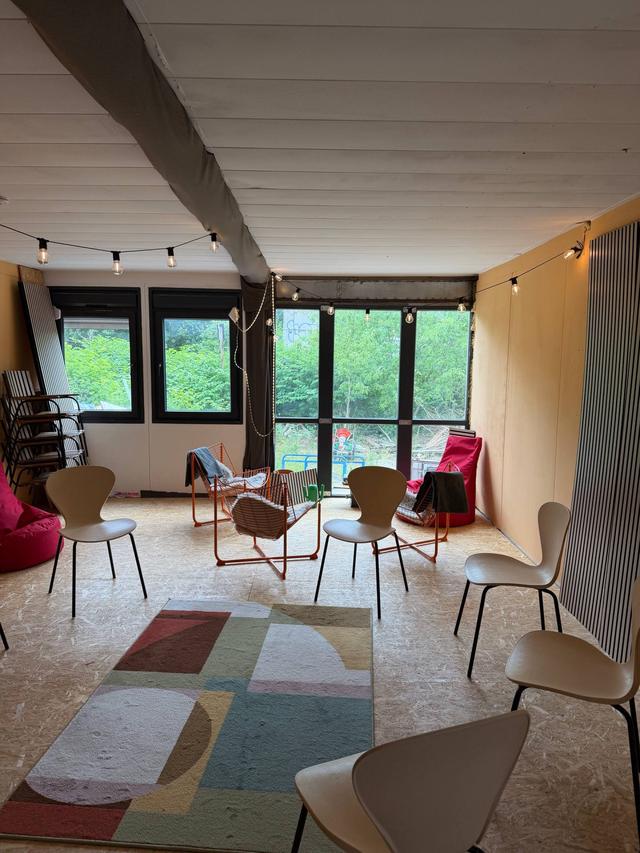 Heller Raum mit Fensterfront zum Grün, Holzstühle, zwei pinke Sitzsäcke, orangefarbene Drahtsessel und ein bunter Teppich, Lichterkette an der Decke