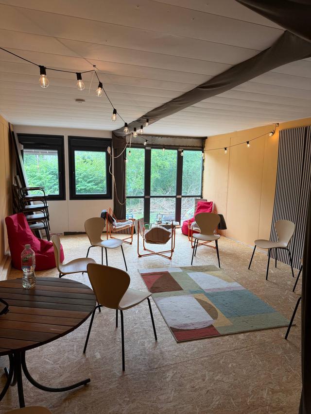 Blick durch den Seminarraum: runder Holztisch, Stühle, bunter Teppich und Lichterkette, hinten die Fensterfront